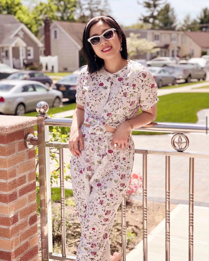 Girl wearing a floral set, sunglasses, pearl headband, with a backdrop of houses and grasses.