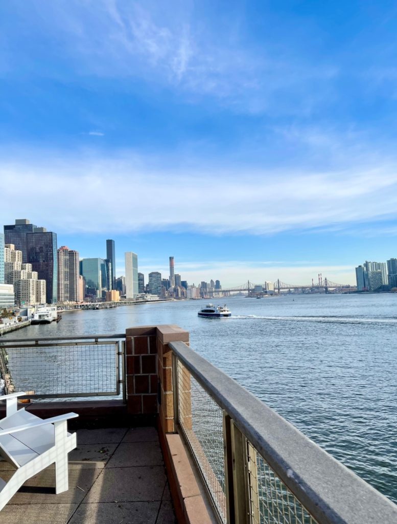 View from a balcony showing the East River and a ferry with a backdrop of New York City buildings.