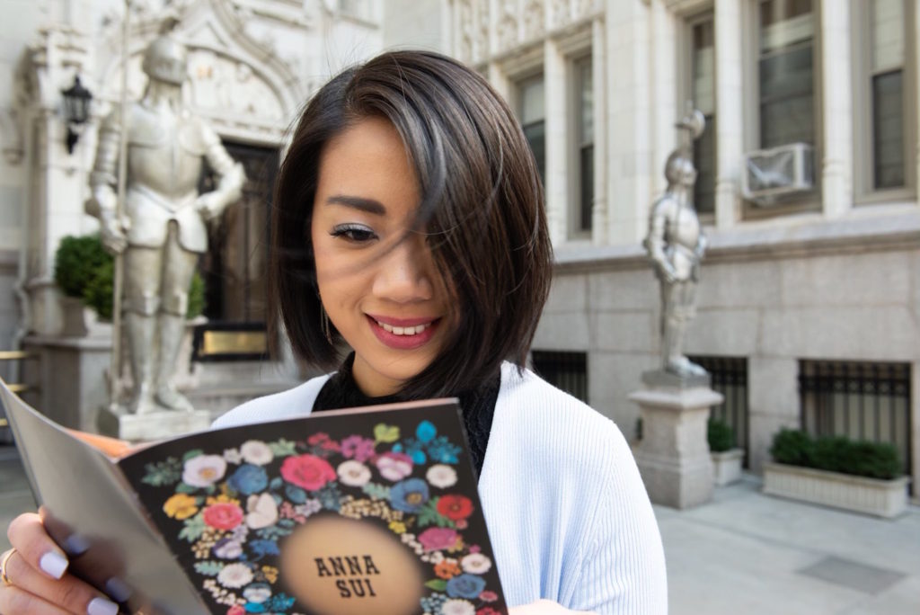 Wendy Sy wearing a blue sweater and holding an Anna Sui Cosmetics brochure in Gramercy Park, New York.