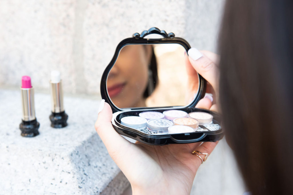 Wendy Sy looking at a reflection of an eyeshadow palette with pink and glitter lipsticks in the background.