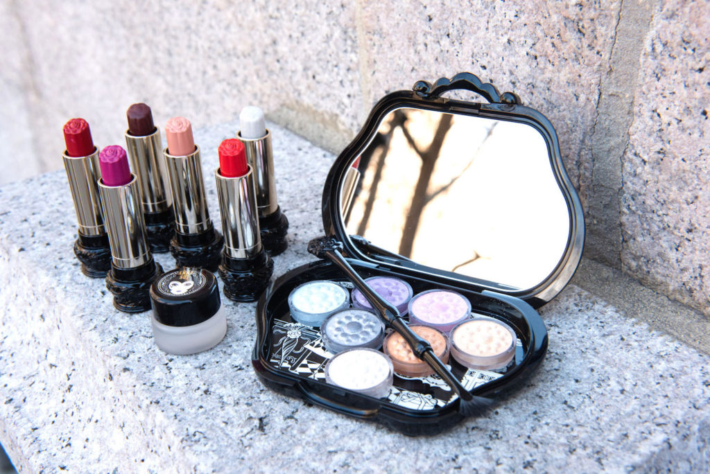 An open eyeshadow palette next to dix lipsticks in various colors by Anna Sui Cosmetics on a gray marble countertop.