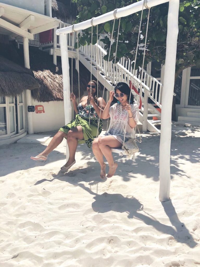 Wendy Sy and Taylor Moon on a swing set with a background of a staircase and sand on a beach in Tulum.