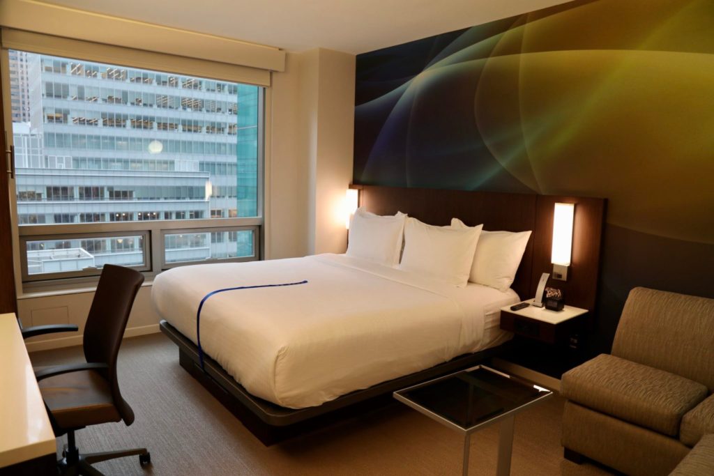 A bed with white sheets next to a window facing New York City during a NYFW staycation at LUMA Hotel Times Square.