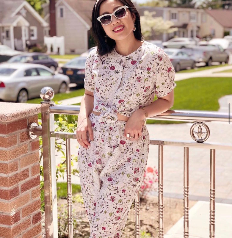 Girl wearing a floral set, sunglasses, pearl headband, with a backdrop of houses and grasses.