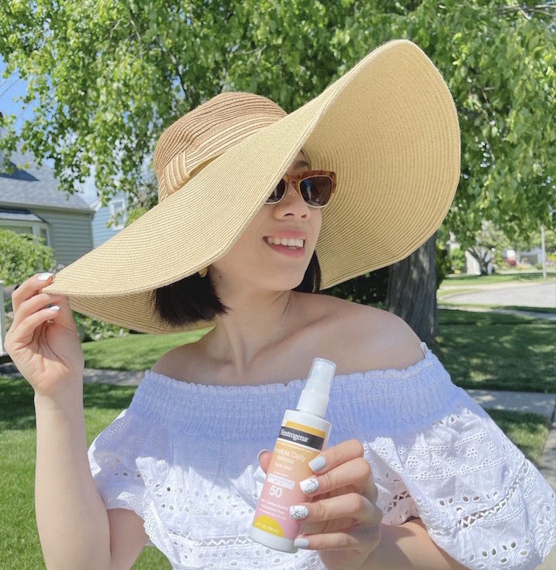 Girl wearing a sunhat holding a Neutrogena sunscreen face mist with a background of trees.