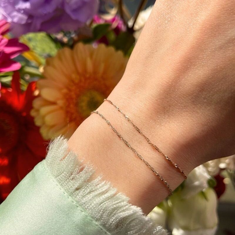 A wrist with two gold permanent jewelry bracelets and a background with colorful flowers.