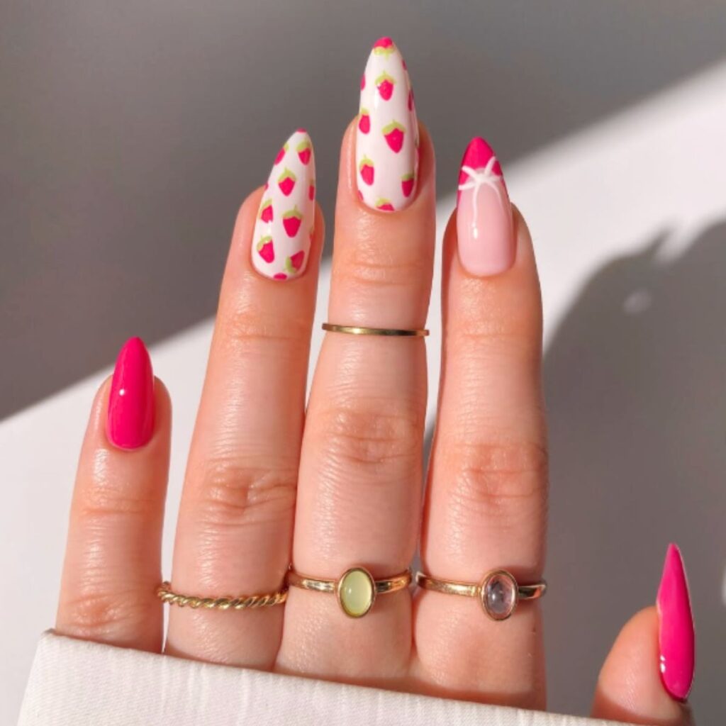 Long pink strawberry nails on a hand with gold rings and a gray background.