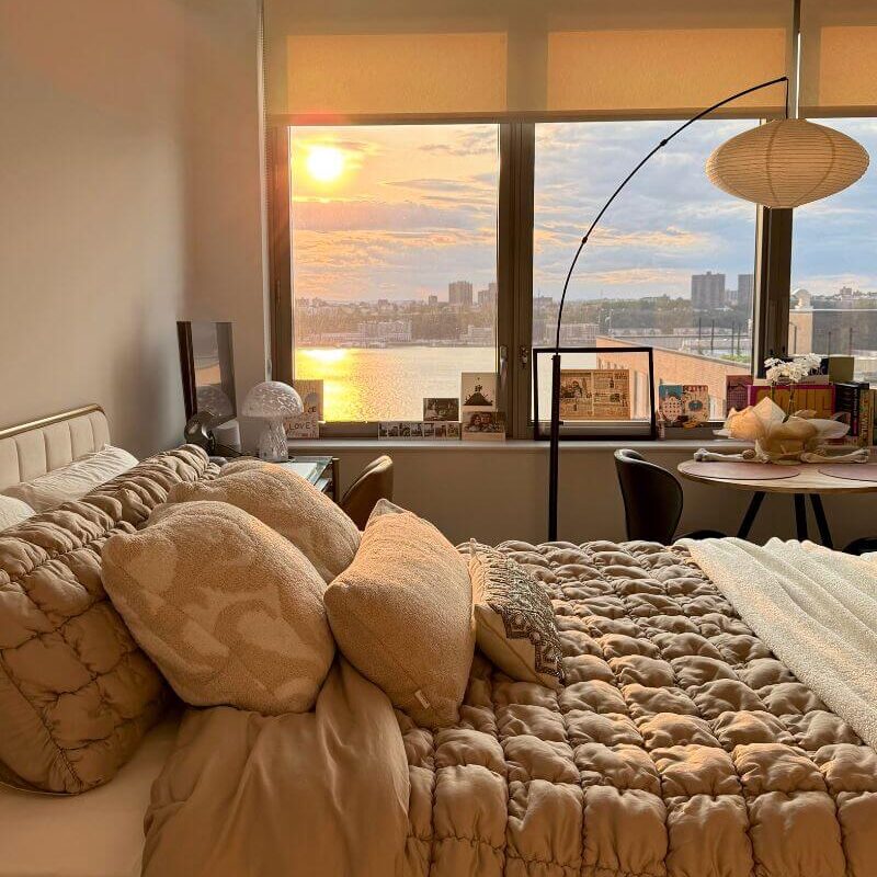 Cozy bedroom with soft lamp light, a bed by the window, and a warm sunset glow.